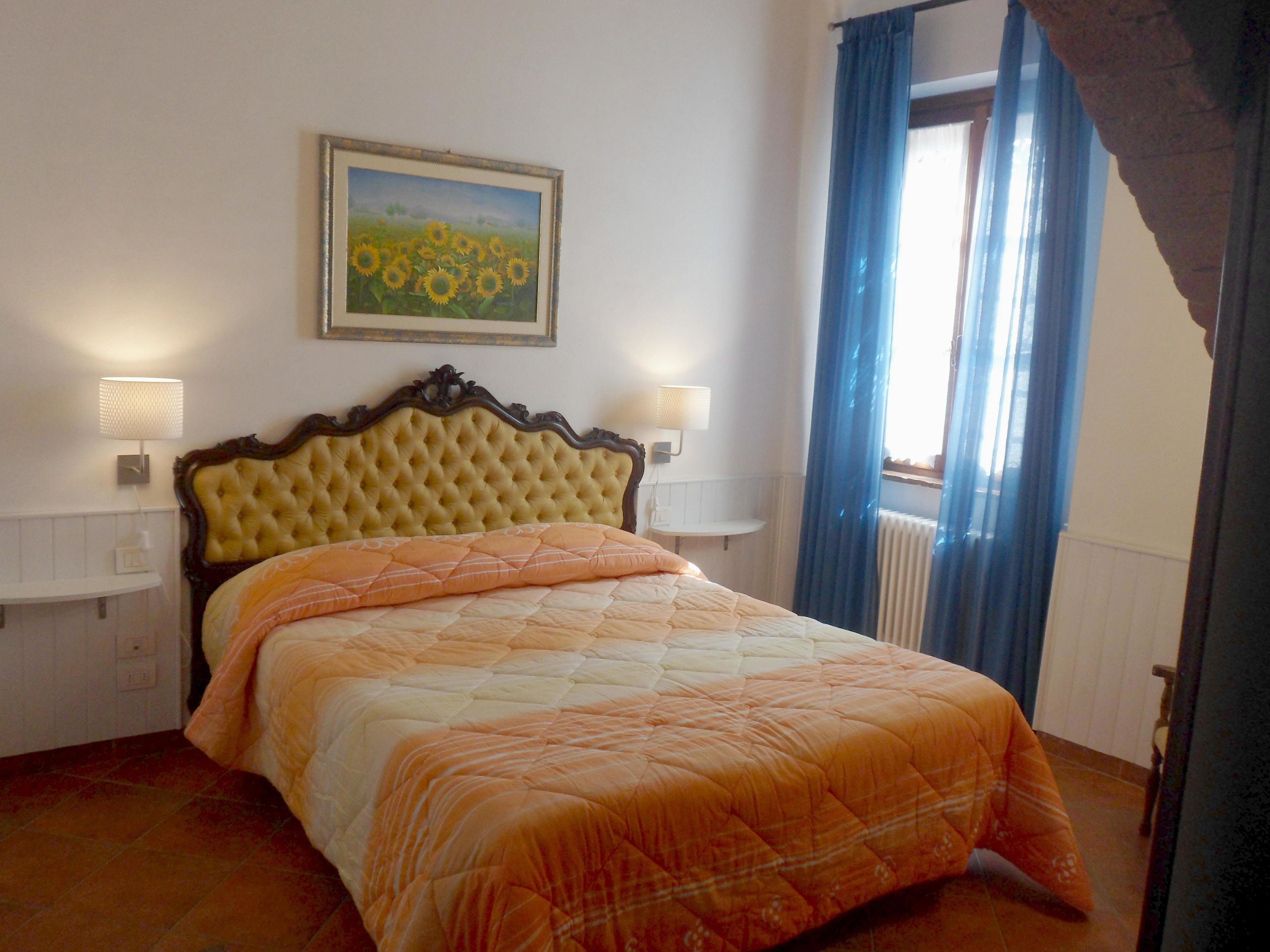 Un dormitorio con una cama grande, dos lámparas de noche, un cuadro enmarcado sobre la cama y una ventana con cortinas.
