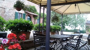 An outdoor patio area with tables and chairs, surrounded by potted plants and a brick building with green shutters. - Agriturismo il Poggiarello (Siena)