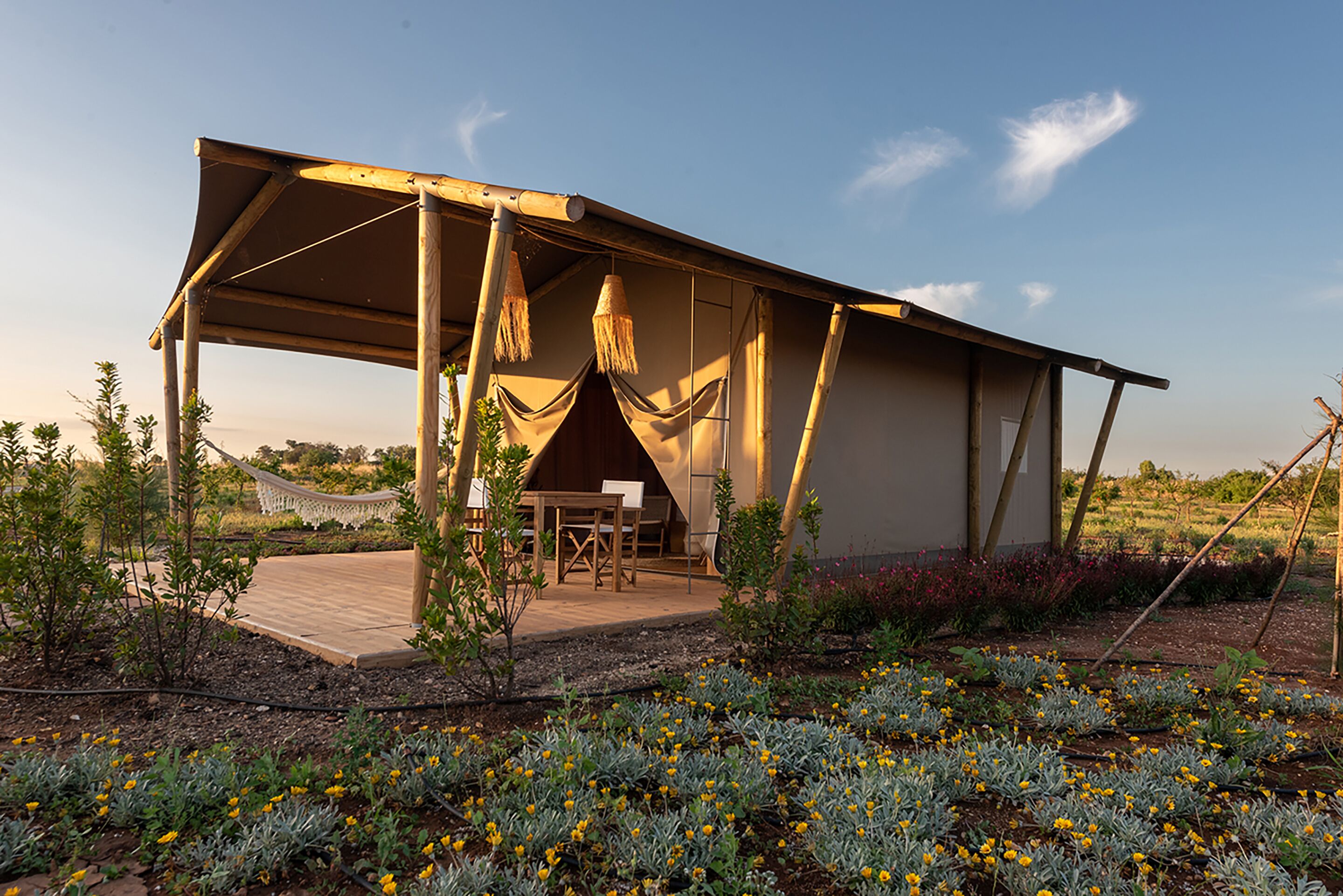 Un área de estar al aire libre techada, con una mesa y sillas, rodeada por un jardín con diversas plantas y flores.