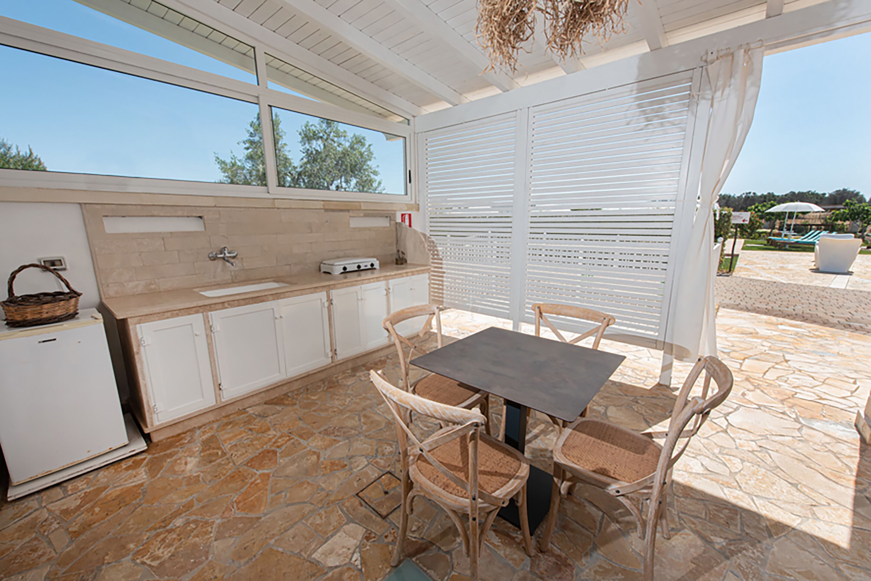 Una cocina con piso de piedra, armarios blancos, una mesa de comedor con sillas y un ventanal con vista a un jardín.