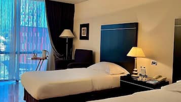 A hotel room with two beds, a desk with a lamp, a chair, and a large window with curtains.