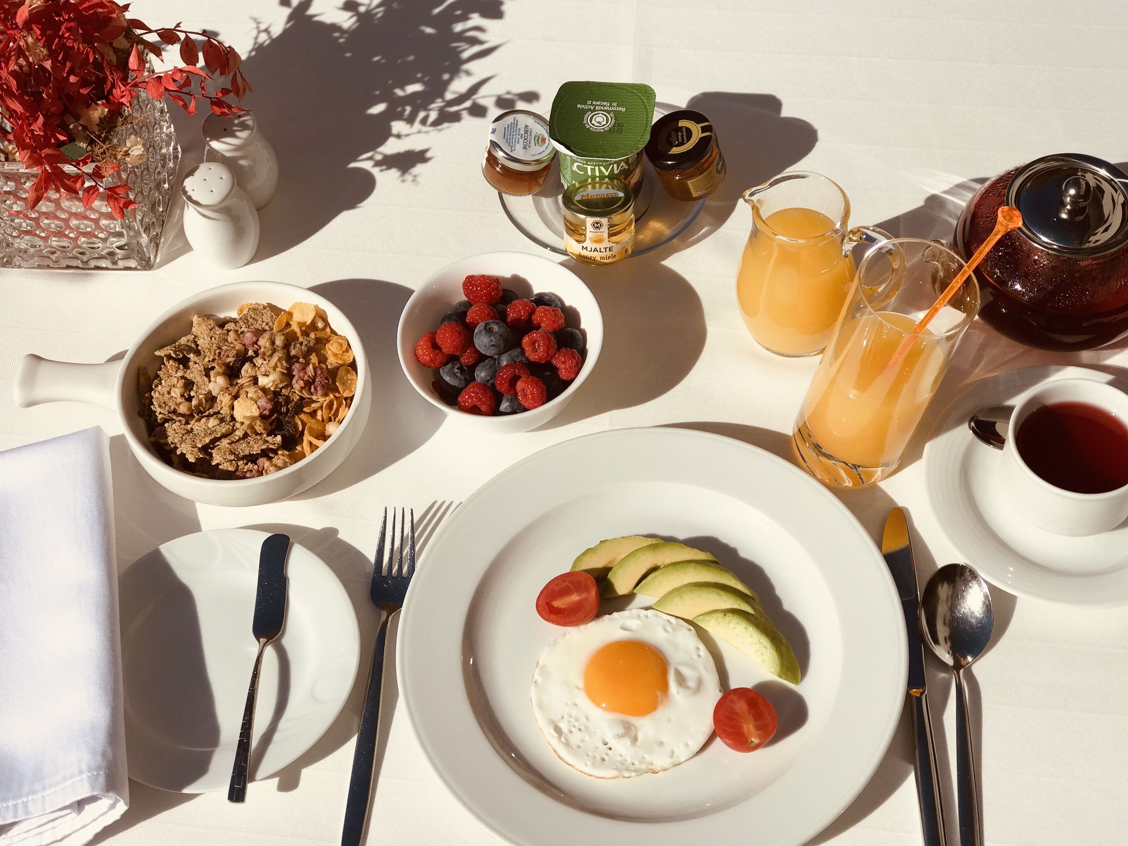 A breakfast table with a plate of avocado and egg, bowls of cereal and berries, honey, and a glass of orange juice.