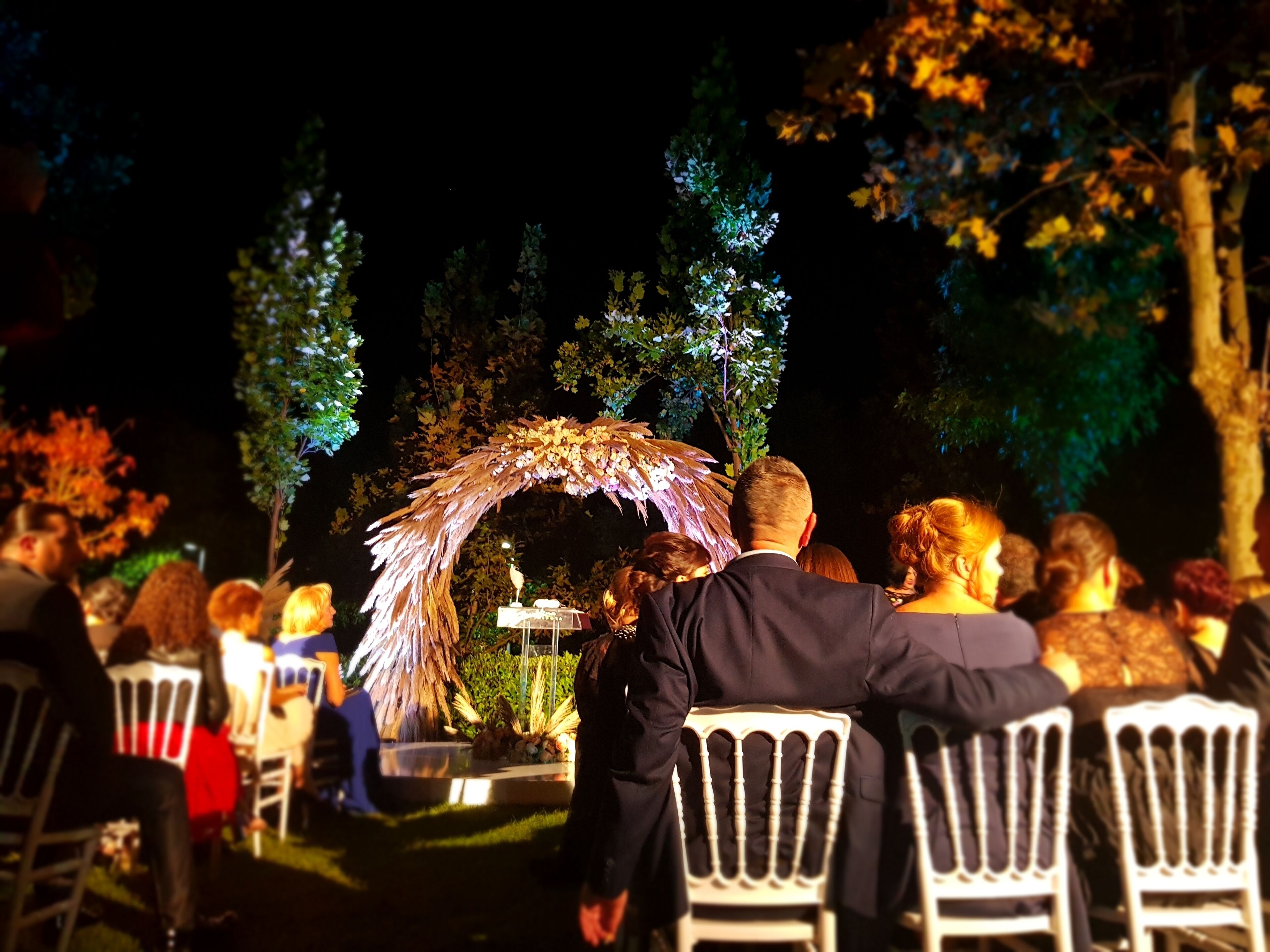 An outdoor event with guests seated at white chairs, an illuminated floral arch, and trees in the background.