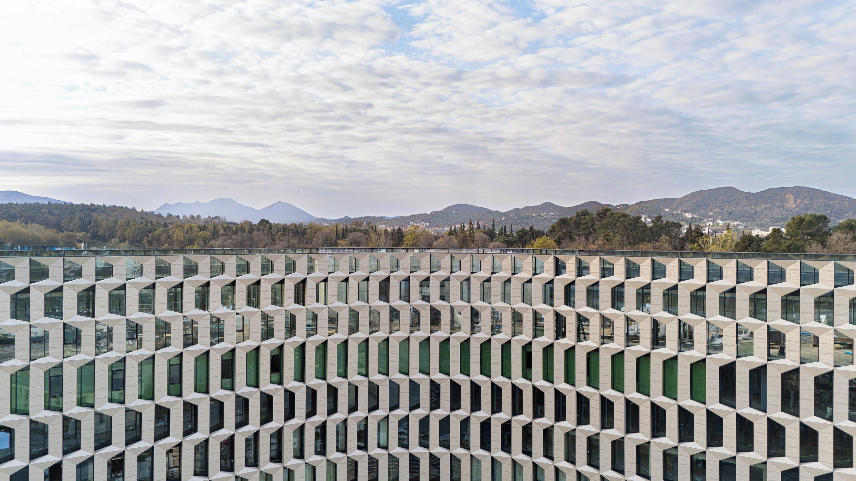 A modern building with a geometric facade and large windows, set against a backdrop of mountains and trees.