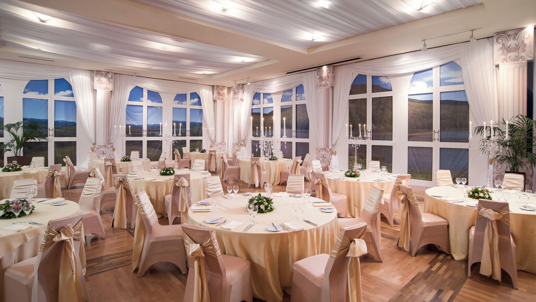 Un salón de banquetes con mesas redondas, sillas y ventanas amplias con cortinas.