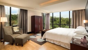 Quarto de hotel moderno com uma cama grande, área de estar com uma poltrona e uma mesa, guarda-roupa e janelas amplas com vista para o exterior.