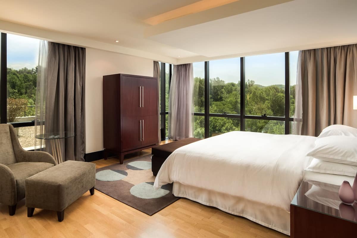 Une chambre d’hôtel moderne dotée d’un grand lit, d’un coin salon avec un fauteuil et une table, d’une armoire et de grandes fenêtres offrant une vue sur l’extérieur.