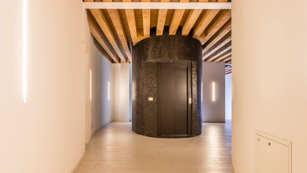 Un hueco de ascensor moderno con diseño circular, rodeado por un pasillo con suelo de madera e iluminación empotrada.
