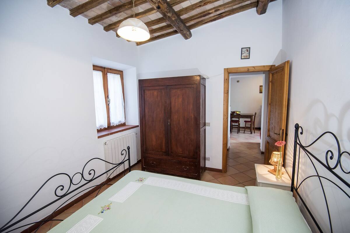 Une chambre à coucher comprenant un lit, une armoire en bois, une fenêtre avec des rideaux, un radiateur et une porte donnant sur une autre pièce.