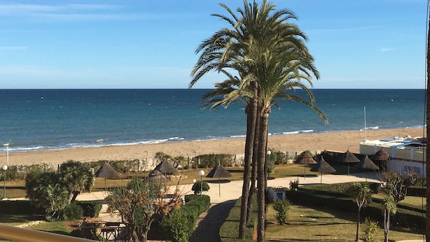 A beach view with a palm tree, beach umbrellas, and a clear sky. - 1ªLínie! Directly on the beach, sea view, pool, air conditioning, WIFI, close to town (Denia)