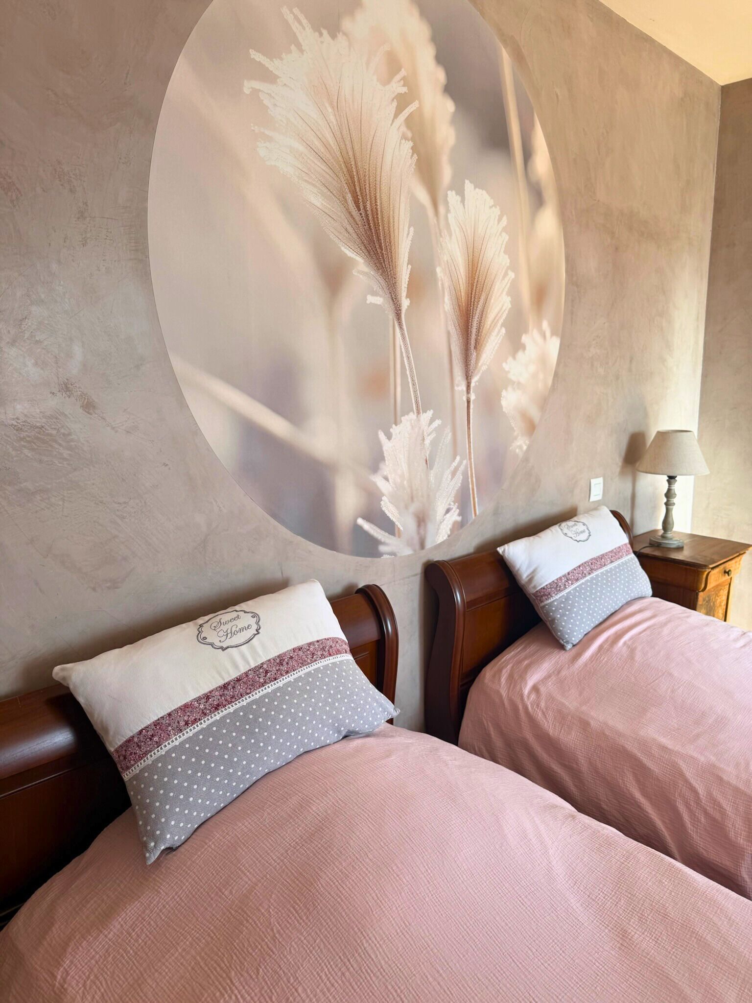 A bedroom with two beds, each with a pink bedspread and matching pillows. There is a wall-mounted artwork of pampas grass and a small bedside table with a lamp.