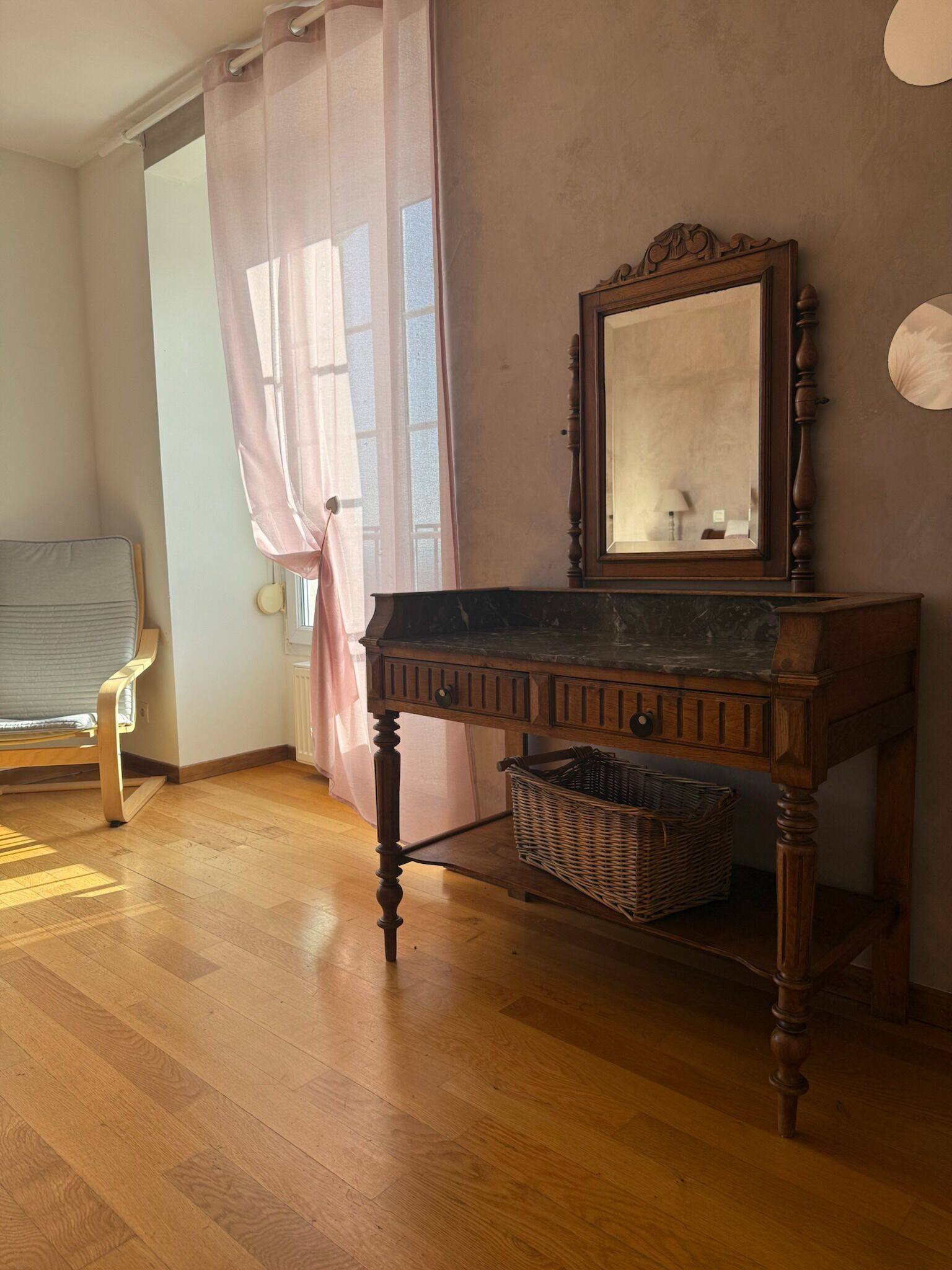A room with a wooden floor, a chair, a table with a mirror, and a window with curtains.