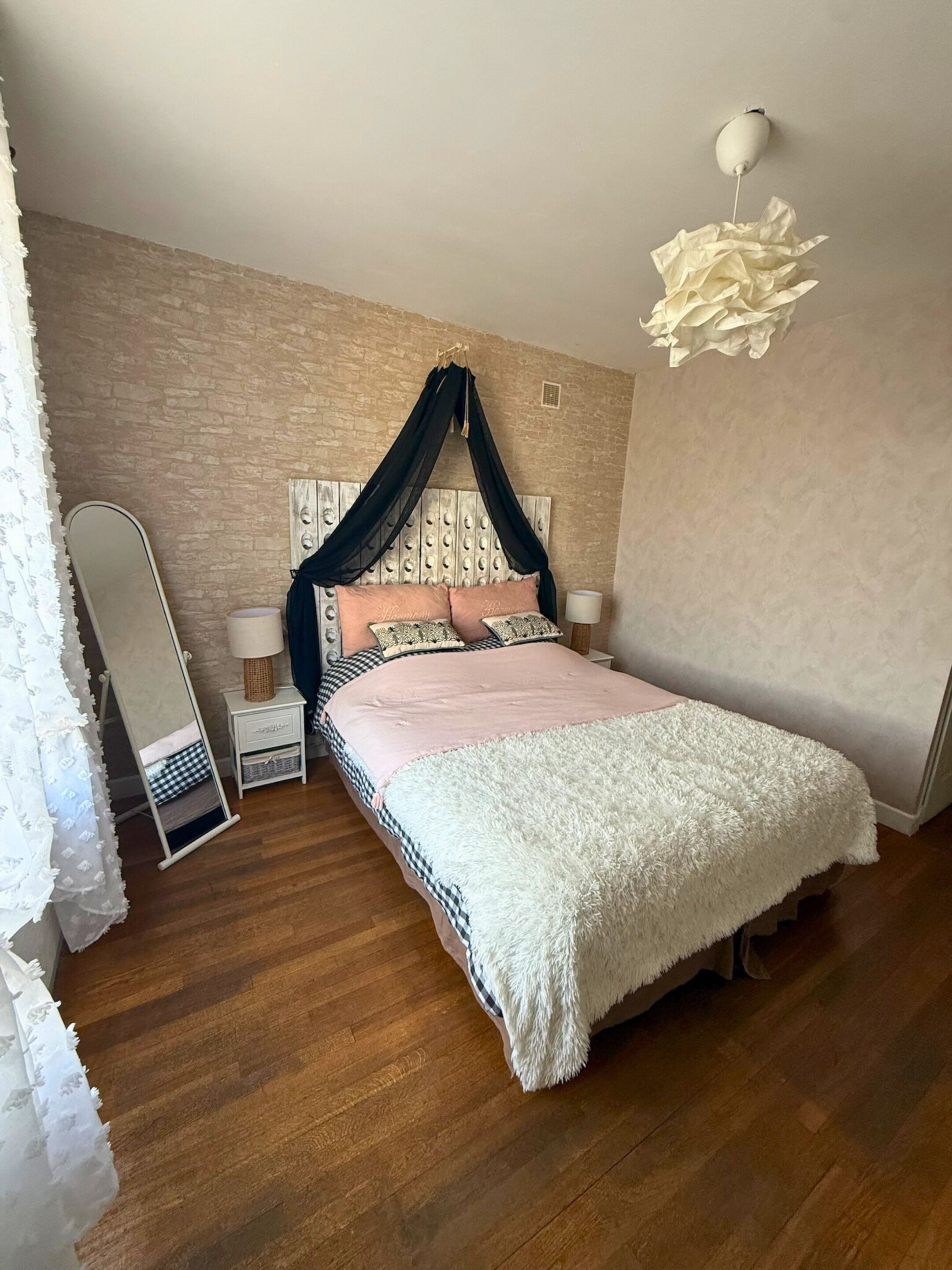 A bedroom with a canopy bed, wooden flooring, a bedside lamp, and a wall-mounted light fixture.
