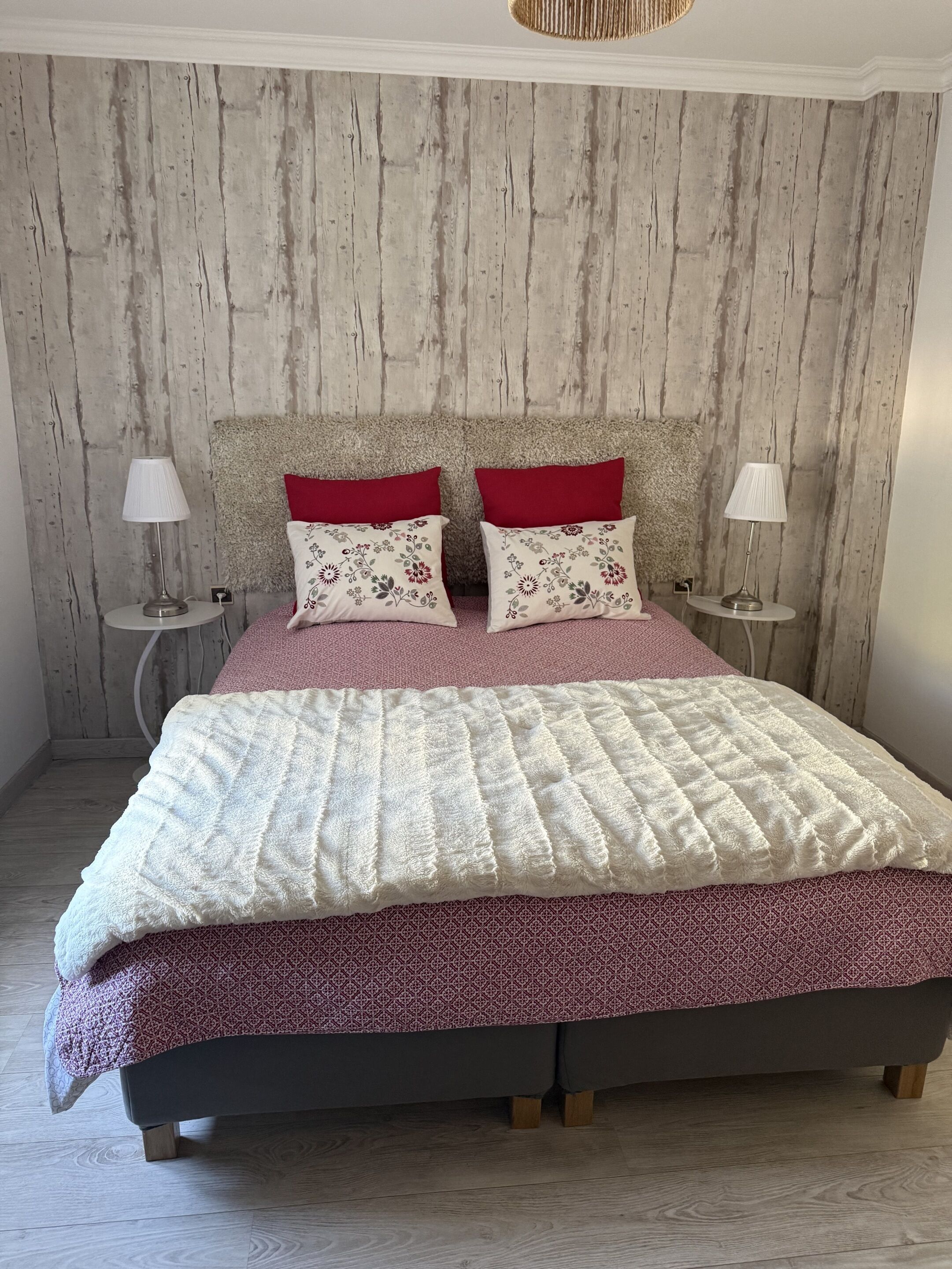 Uma cama com colcha estampada, duas almofadas vermelhas e cabeceira apoiada em uma parede de madeira.
