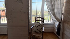 A room with a wooden floor, a chair with a polka dot cushion, a window with a view of a landscape, and a wall with a sketched figure. - Voirin Valérie (Cramant)