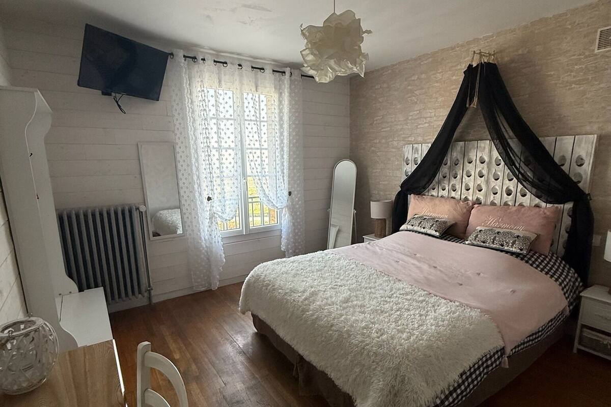 Une chambre à coucher avec un lit à baldaquin, un plancher en bois, un radiateur blanc, un téléviseur fixé au mur et une fenêtre avec des rideaux.