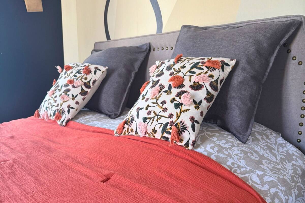 Een bed met een rode deken, grijze kussens en kussens met een bloemenmotief.