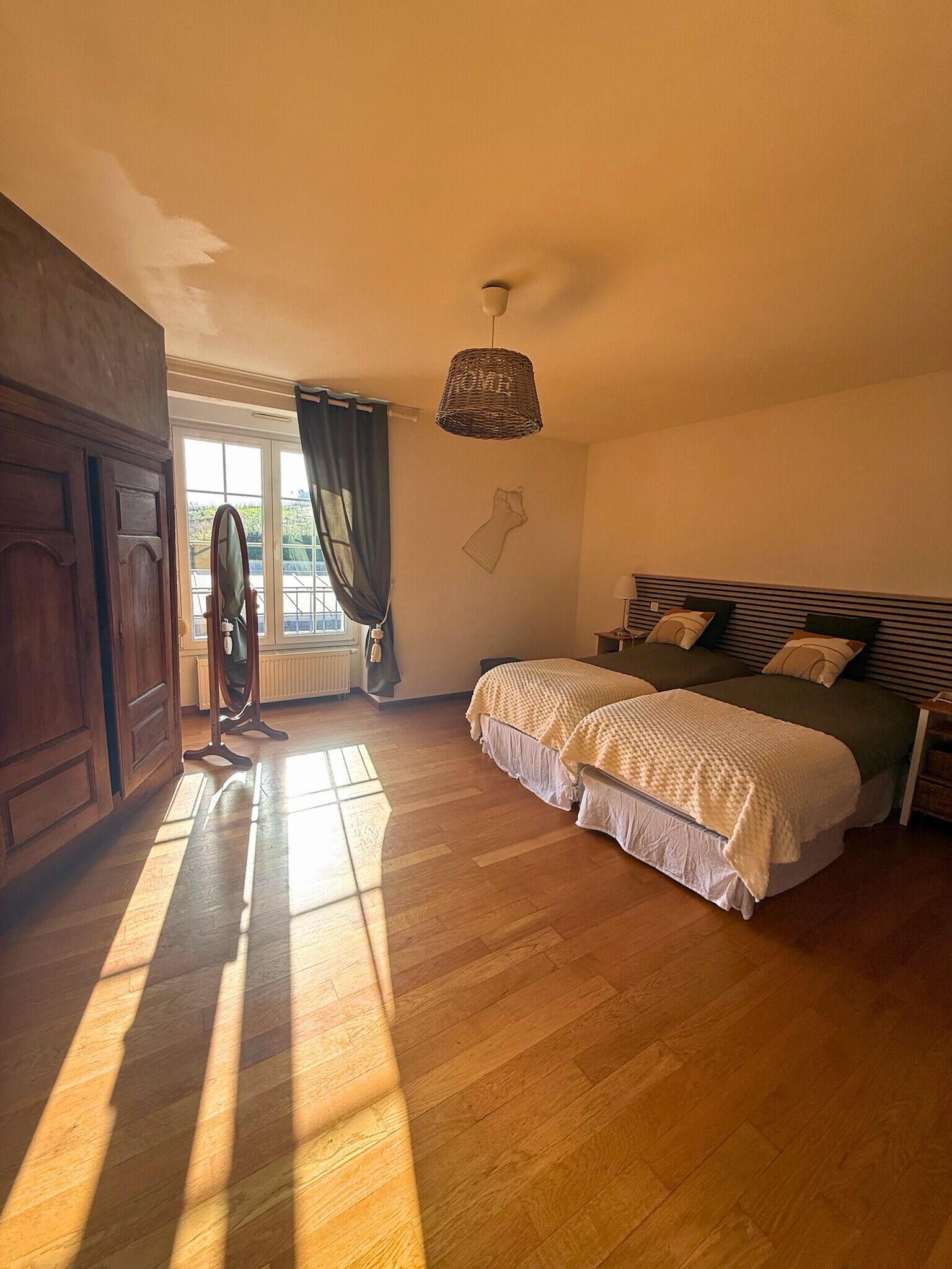 Um quarto com piso de madeira, cama com edredom verde e branco, luminária pendente e janela com cortinas.