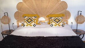 A bed with a rattan headboard, two patterned pillows, a black furry rug, and two round wall lamps. - Voirin Valérie (Cramant)