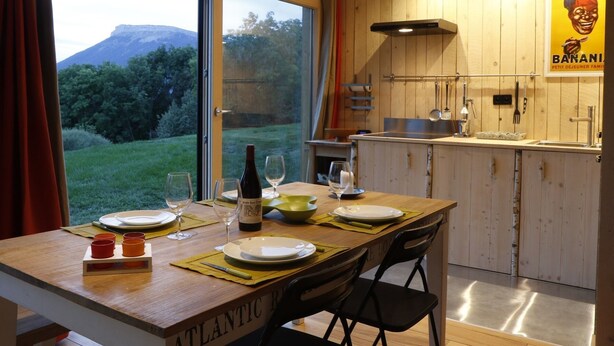 Un comedor con una mesa preparada para cuatro, una cocina con armarios de madera y vistas a un paisaje verde a través del ventanal.
