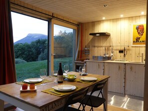 Un comedor con una mesa preparada para cuatro, una cocina con armarios de madera y vistas a un paisaje verde a través del ventanal.