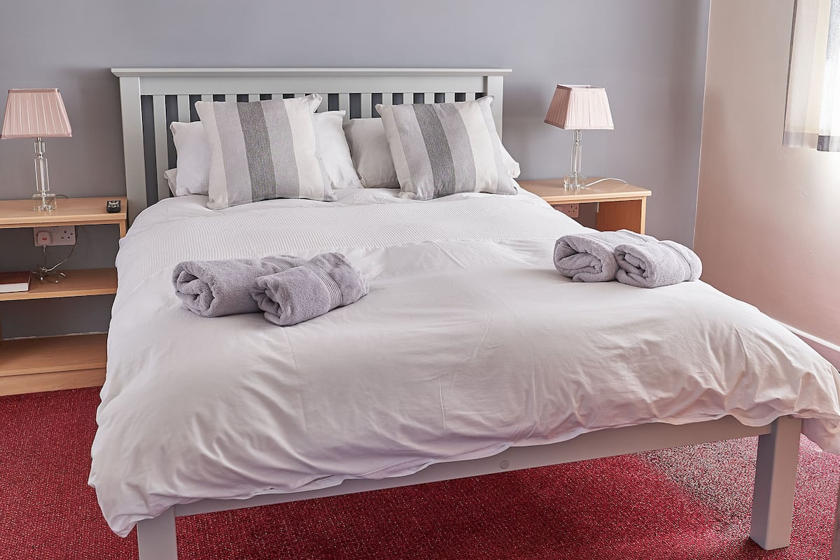 Ein ordentlich bezogenes Bett mit weißer Bettwäsche und zwei Kissen, ein roter Teppich sowie zwei Nachttische mit Nachttischlampen.