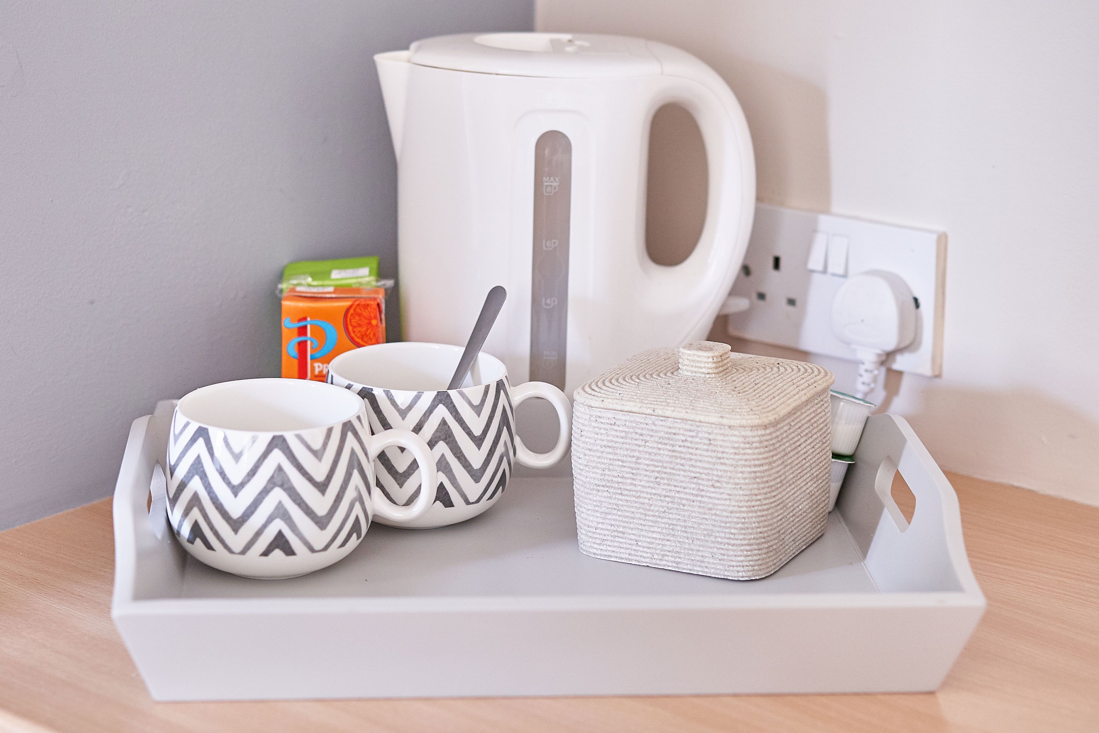 A tray with a white kettle, two patterned mugs, a box of orange juice, and a woven container on a wooden surface.