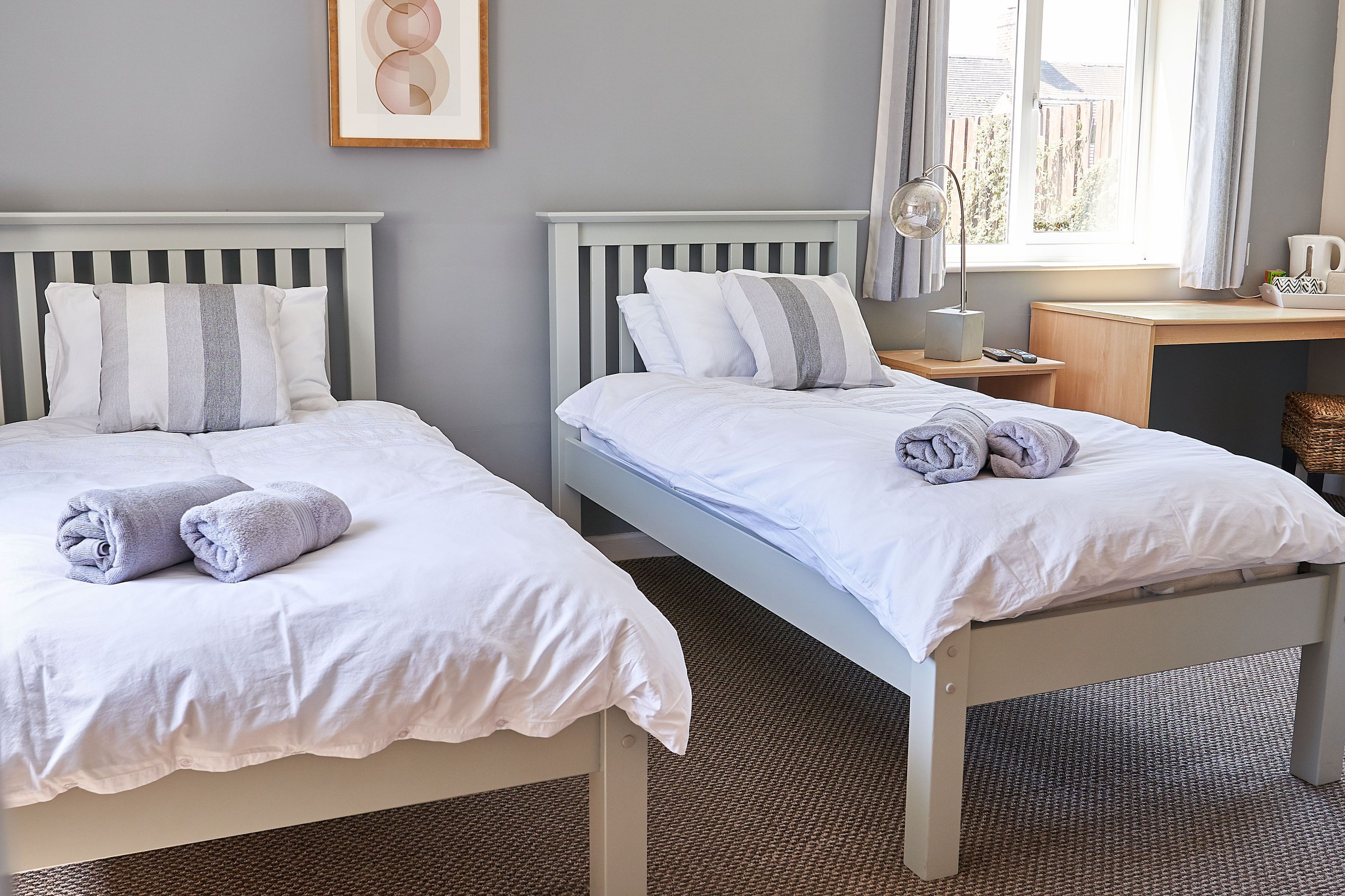Dos camas individuales con ropa blanca y almohadas a rayas, una mesita de noche con lámpara, y una ventana con cortinas.