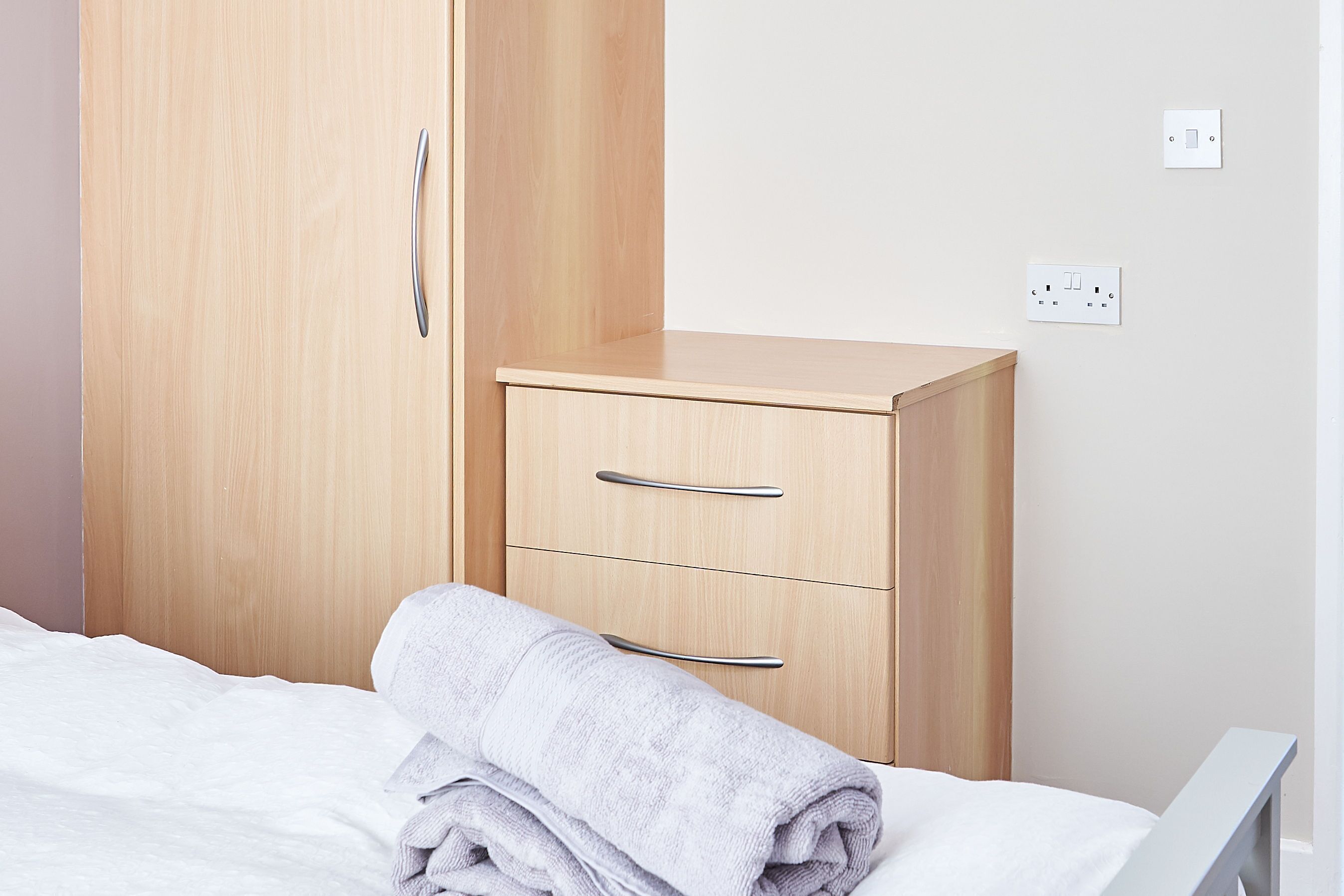A neatly made bed with a folded towel, a wooden wardrobe with two drawers, and a wall socket.