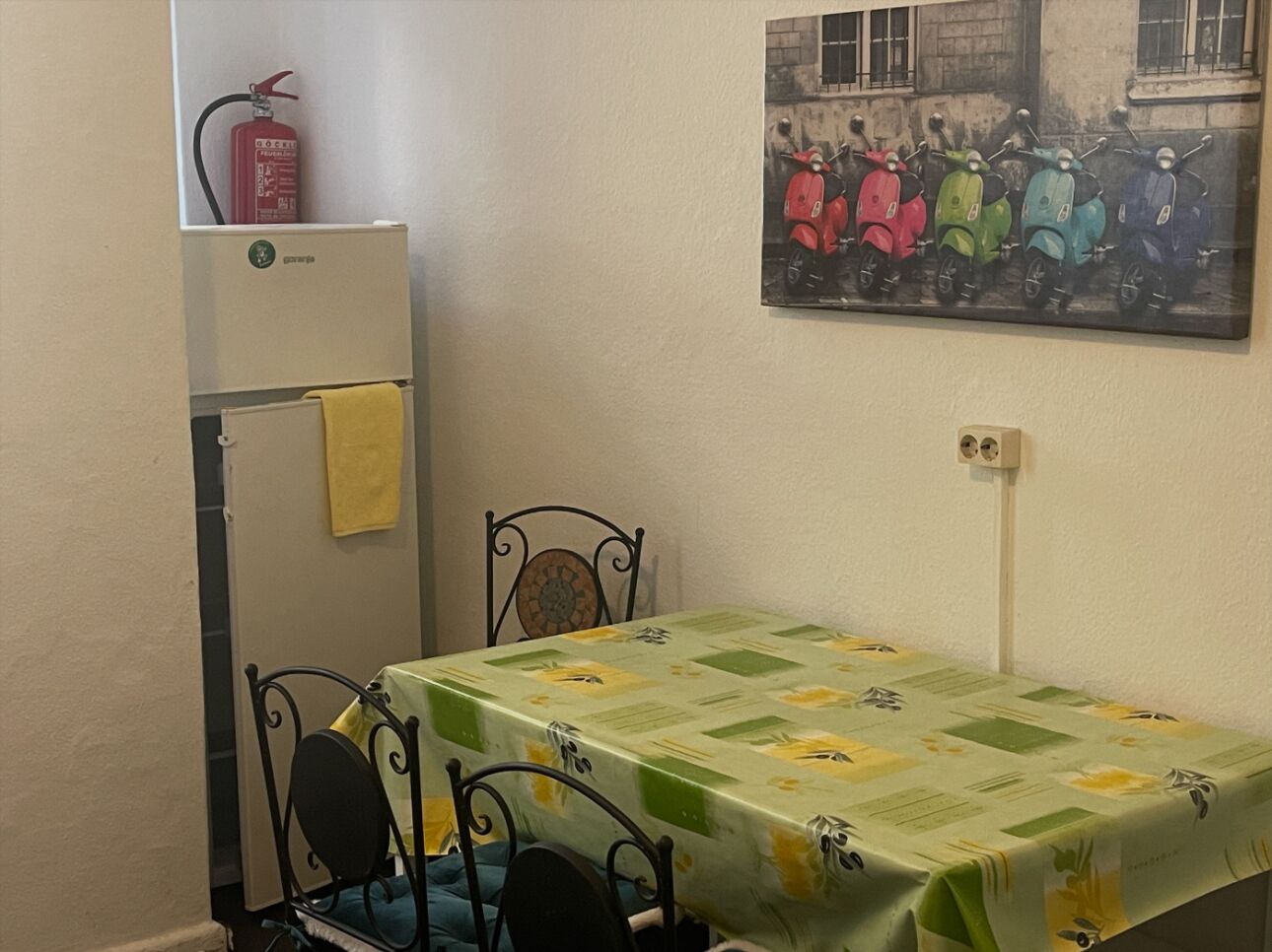 A small room with a table and chairs, a fire extinguisher, a refrigerator, and a painting on the wall.
