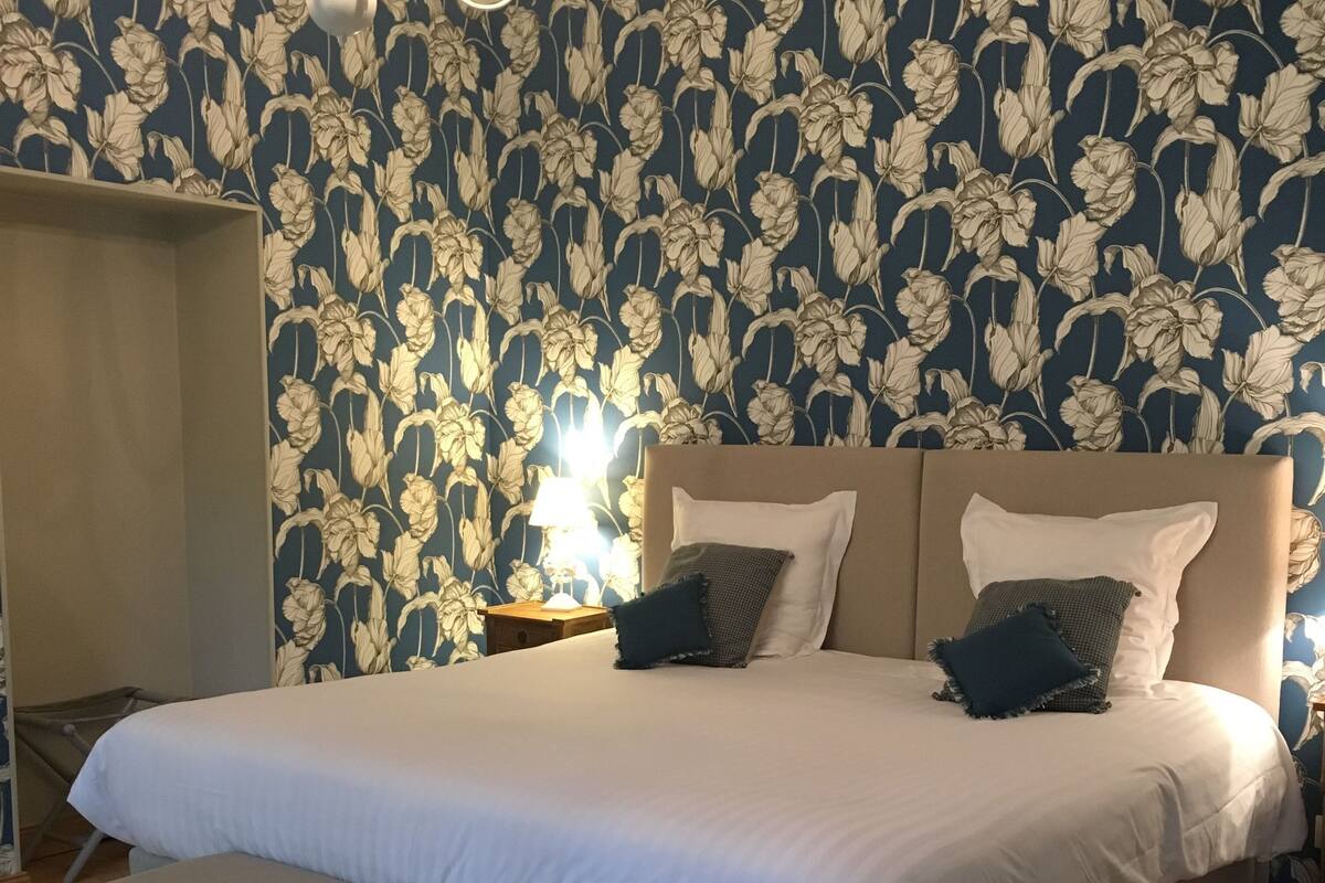 A bedroom with a floral wallpaper, a bed with white bedding, two pillows, a nightstand with a lamp, and a chandelier.
