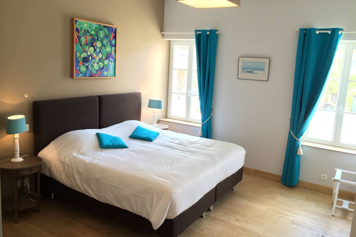 A bedroom with a large bed, two bedside lamps, a window with curtains, a painting on the wall, and a small table.