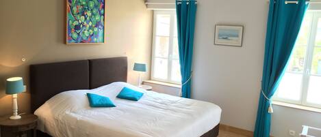 A bedroom with a large bed, two bedside lamps, a window with curtains, a painting on the wall, and a small table.
