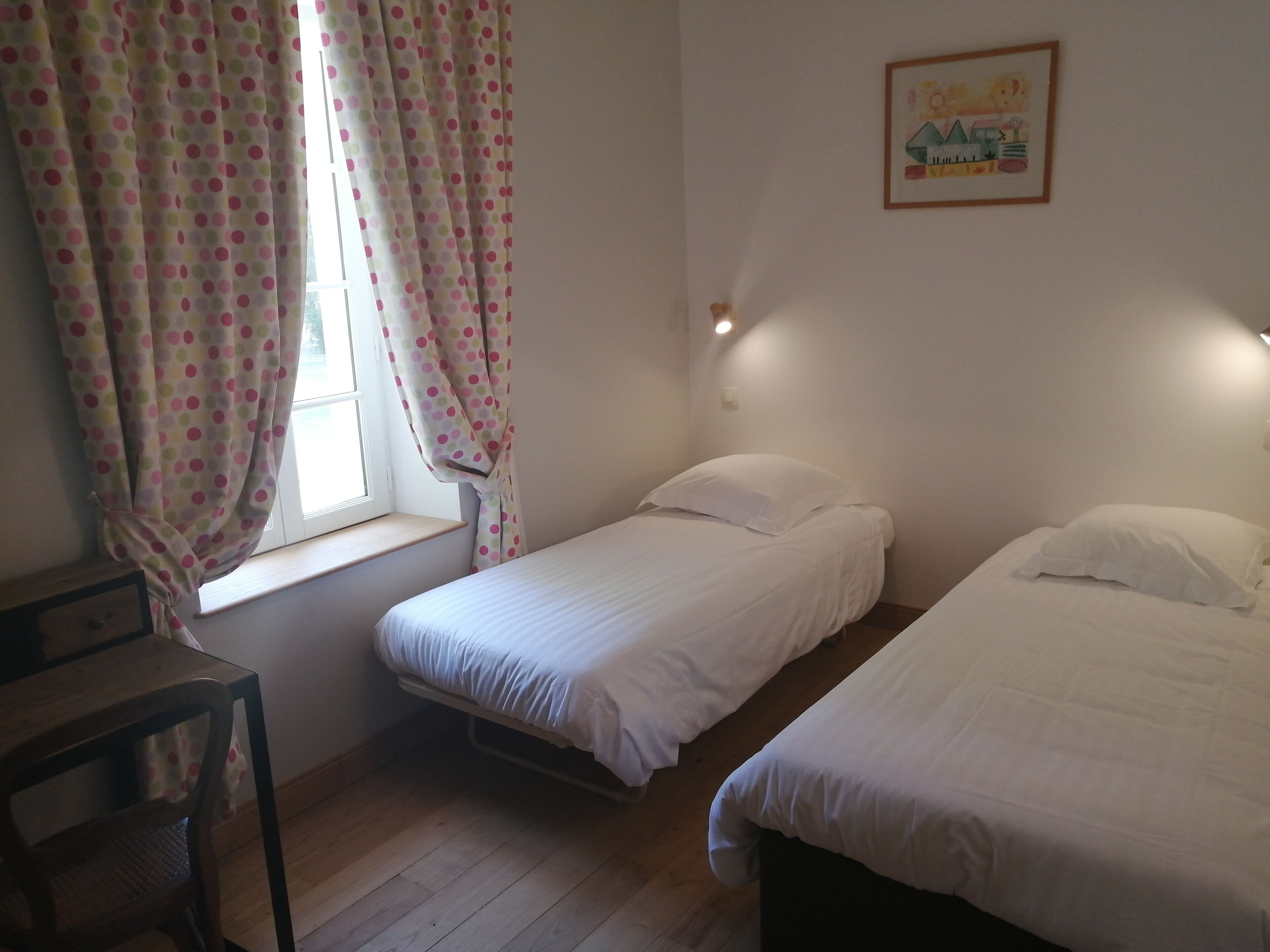 A room with two single beds, a window with curtains, a wooden chair, and a wall-mounted light fixture.
