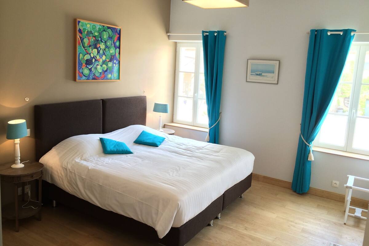 A bedroom with a bed, two bedside lamps, a window with curtains, a painting on the wall, and a small table.