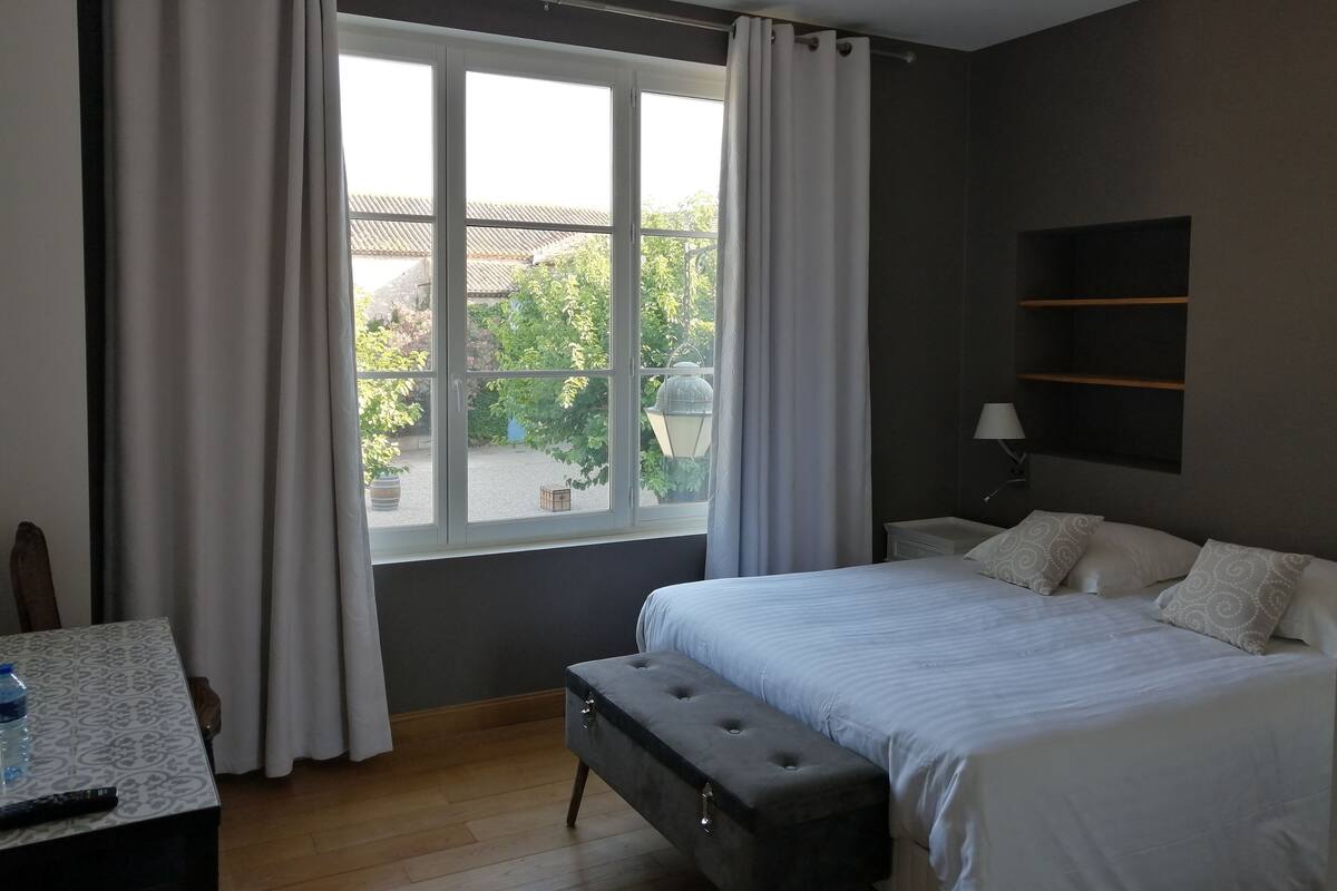 A bedroom with a large window, a bed, a bench, a bedside table, and a lamp.