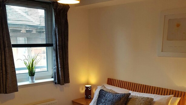 Ein ordentlich bezogenes Bett mit gestreifter Bettdecke, Kissen und zwei gefalteten Handtüchern. Eine Nachttischlampe, ein Fenster mit Vorhängen und ein kleiner Blumentopf auf der Fensterbank sind ebenfalls zu sehen.