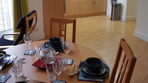 Ein Essbereich mit einem runden Tisch, gedeckt für vier Personen, einer Küche im Hintergrund und Blick ins Freie durch ein Fenster mit grünen Vorhängen.