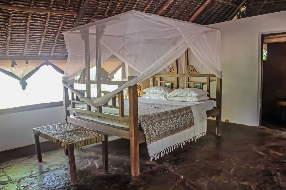 A room with a bed under a white canopy, a wooden bench, and a stone floor.