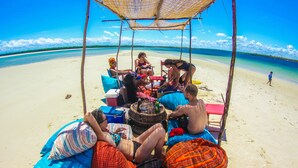 A group of people relaxing under a thatched roof on a sandy beach with clear blue water in the background.
