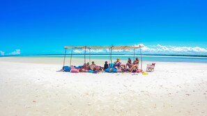 A beach scene with a group of people under a shaded area, a few chairs, and a clear view of the ocean. - Mikoko Cove Ecolodge (Funzi Island)