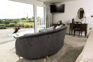 A modern living room with a grey sofa, a flat-screen TV, a desk with a mirror, and a view of the countryside through large windows.