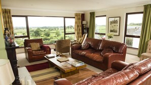 A spacious living room with leather furniture, large windows offering a view of the countryside, and a wooden coffee table with books and decorative items. - Peartree Hill (Belfast)