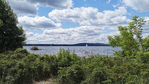 Ein Strand mit dichter Vegetation im Vordergrund, ein sandiger Uferstreifen und ein Blick auf einen See mit Segelbooten in der Ferne.
