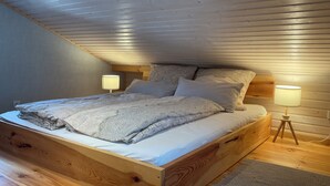 Ein Schlafzimmer mit schrägem Dach, einem Bettgestell aus Holz, gemusterten Betttextilien, zwei Nachttischlampen und einem Teppich mit Fischgrätmuster.