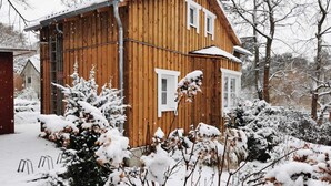 Ein hölzernes Haus mit geneigtem Dach und weißer Verzierung, umgeben von mit Schnee bedeckten Bäumen und Sträuchern.