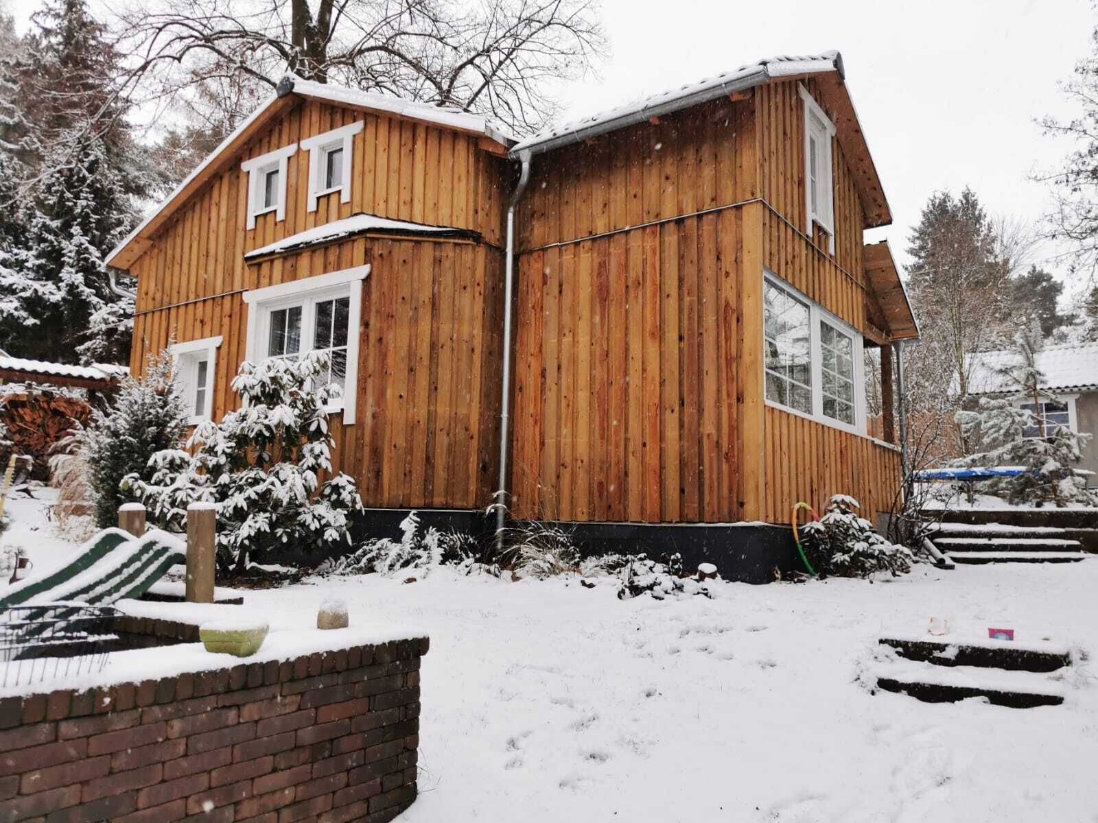 Uma cabana de madeira com telhado inclinado, base de tijolos e quintal coberto de neve.
