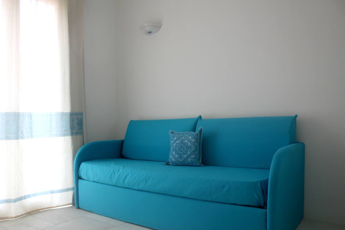 A teal sofa with a patterned pillow against a plain wall, a ceiling light fixture, and a window with a blue-striped curtain.