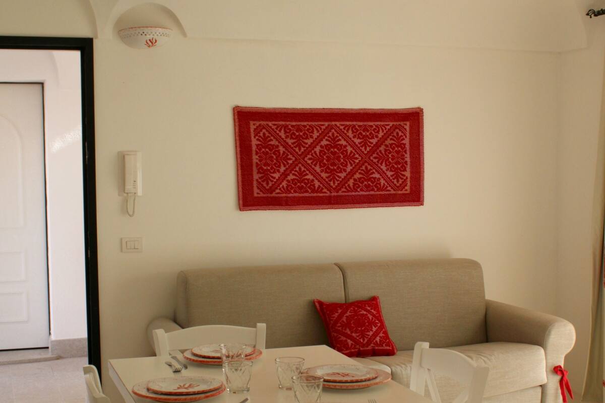 A dining area with a white table set for four, white chairs, a beige sofa, and a red decorative piece on the wall.