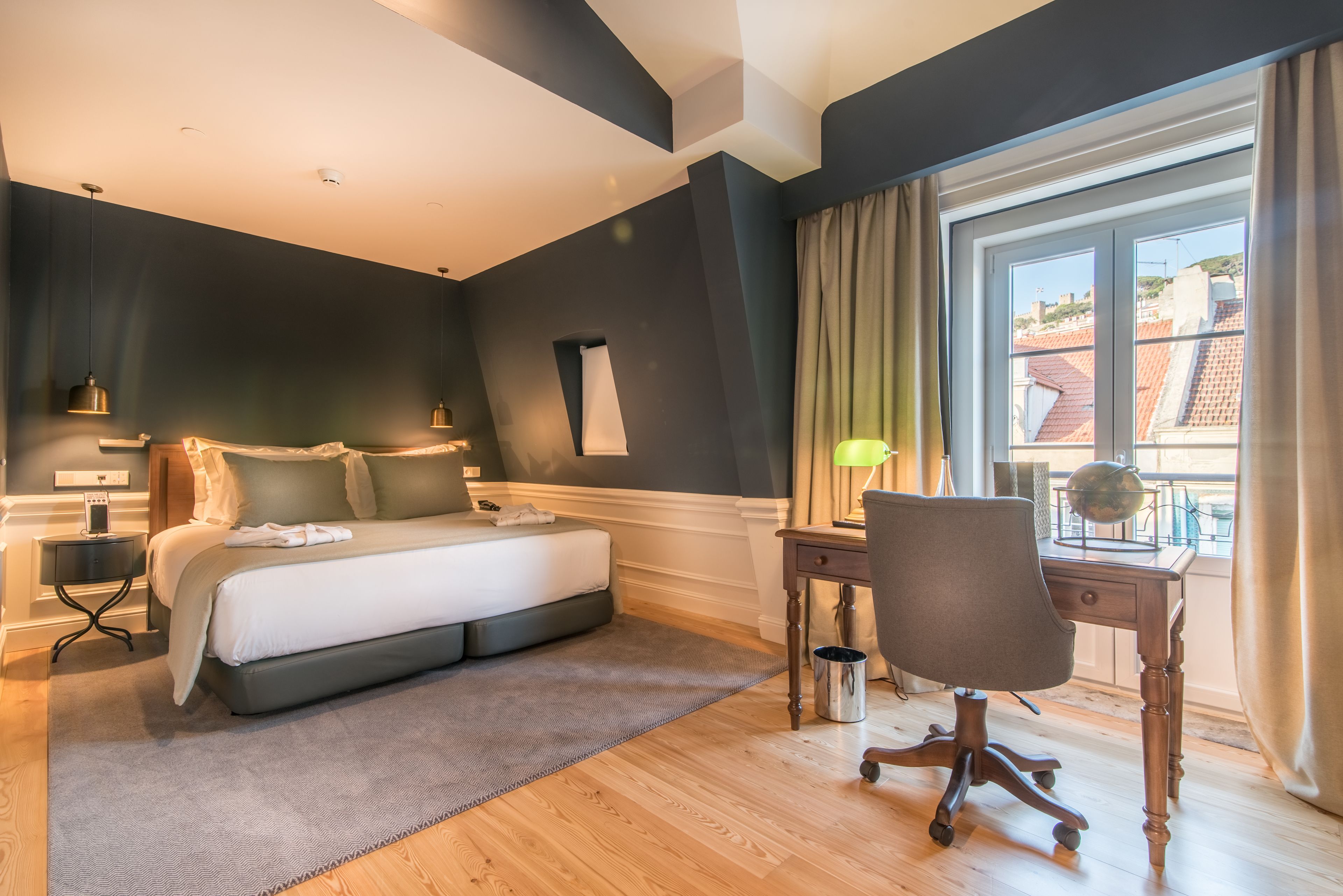 Une chambre à coucher comprenant un lit, des tables de chevet, un bureau avec une chaise, une fenêtre avec des rideaux et une vue sur les bâtiments à l’extérieur.